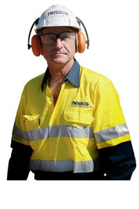 Smaller A man wearing yellow and blue NPS high-visibility work wear, safety hat, glasses and ear protection looks at the camera.